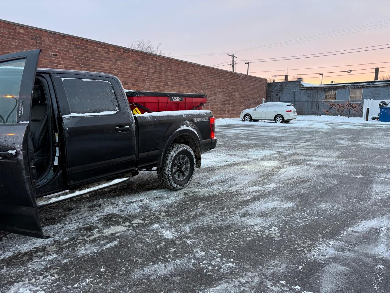 Black pickup truck with a red bulk salt spreader in a treated commercial parking lot at sunrise
